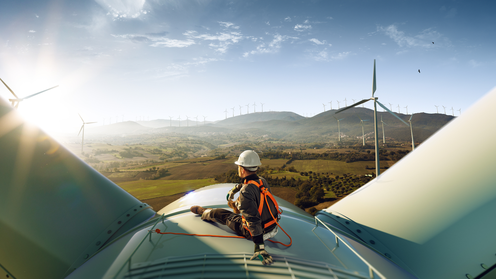 Man in hard hat and safety harness sits on top of a wind turbine overlooking beautiful sunset landscape dotted with other wind turbines