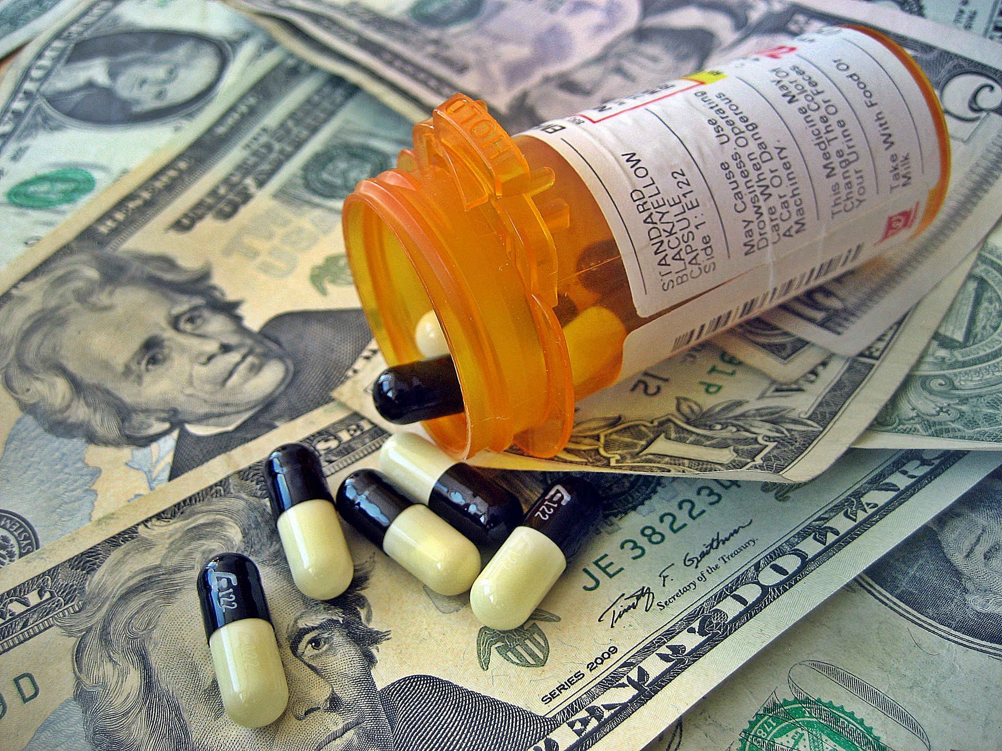 An open prescription bottle spills pills onto twenty dollar bills.