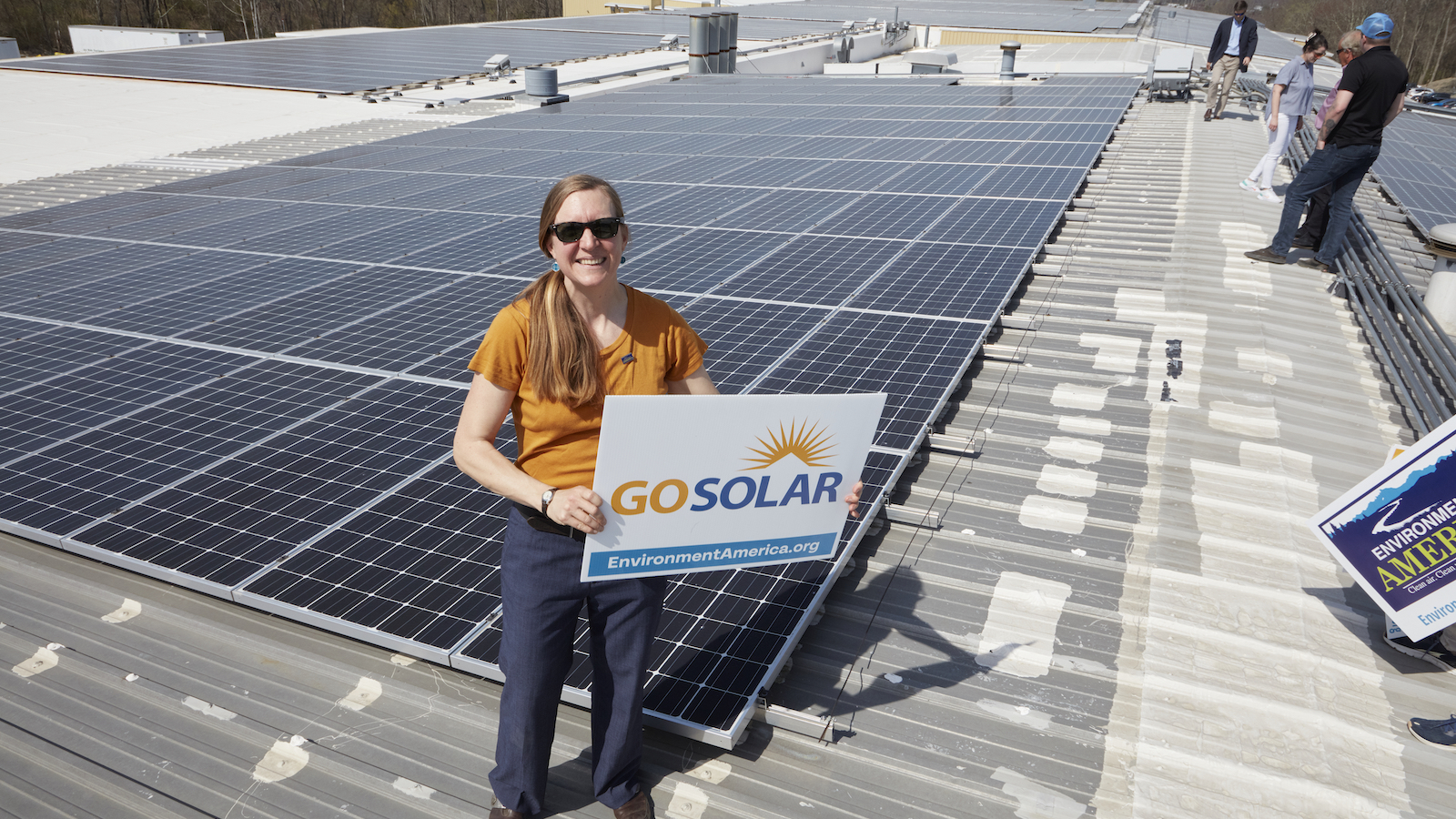 Environment America 100% Renewable Energy Senior Director Johanna Neumann stands on a warehouse rooftop covered in solar panels holding a sign that reads 