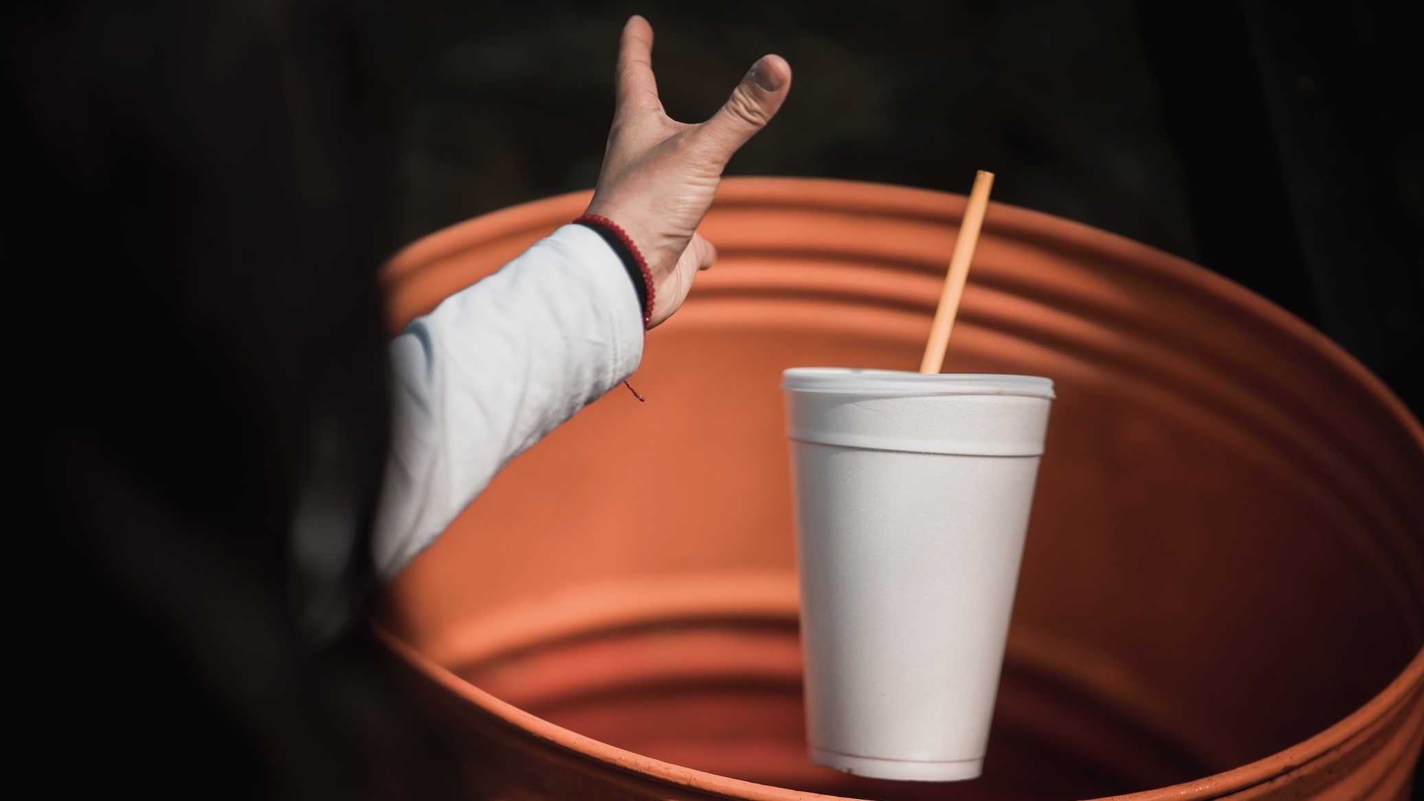 A person throwing away a polystyrene foam cup and straw into an orange bin