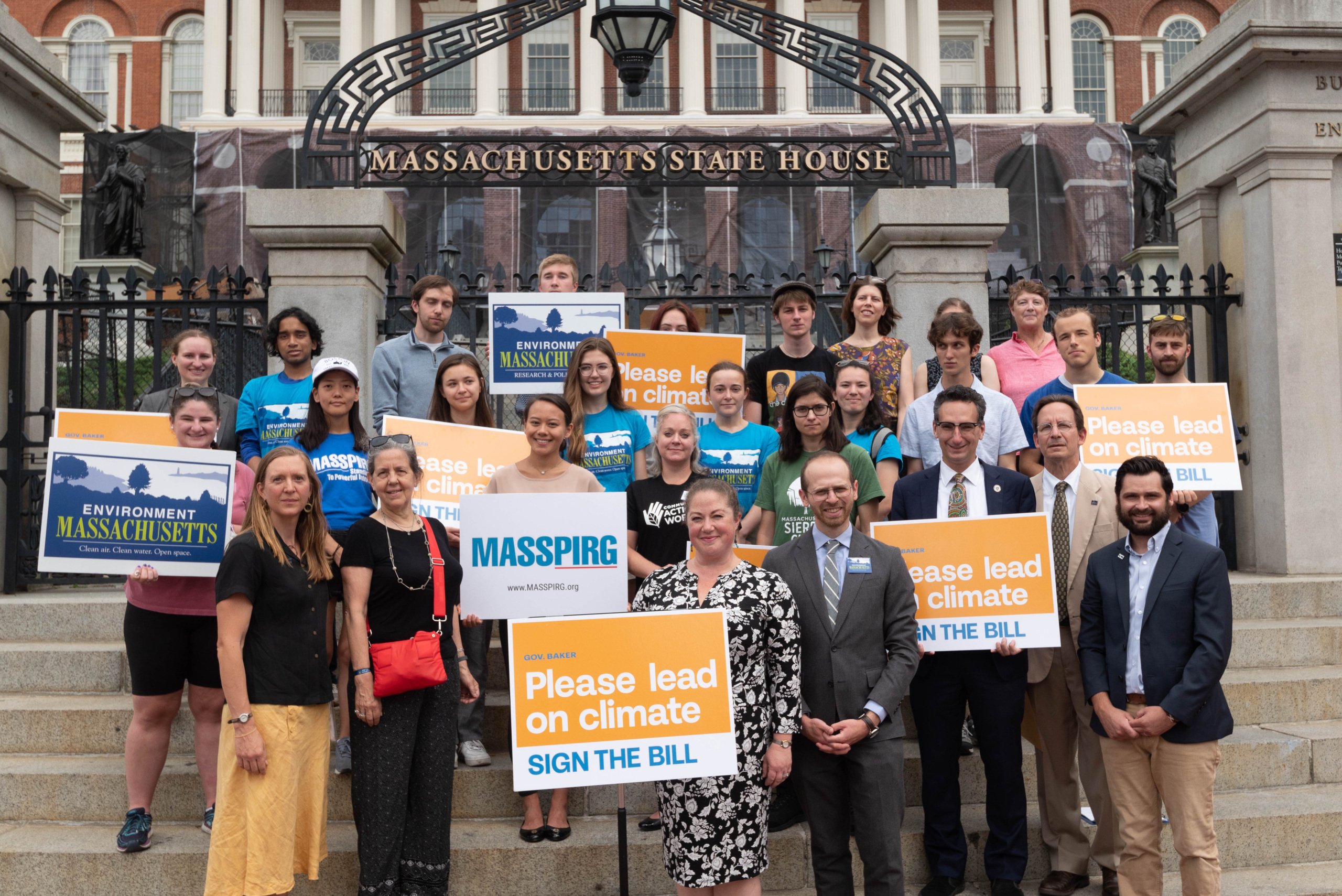 Activists and elected officials stand in front of the Massachusetts State House holding signs asking Gov. Charlie Baker to sign a climate bill.