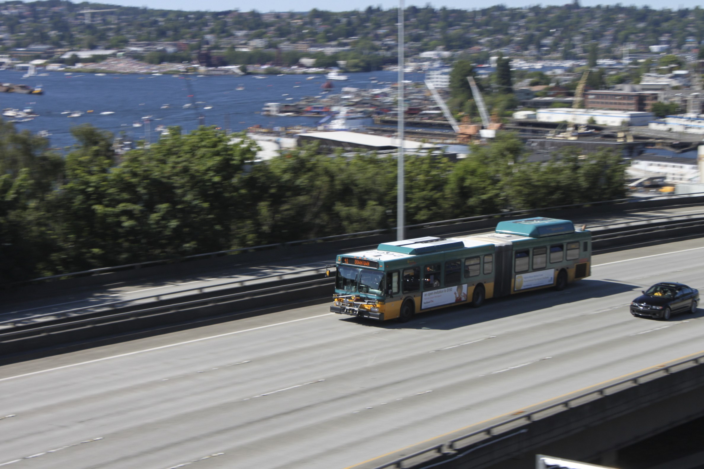 An extended bus drives on an empty freeway near a bay on a sunny day.