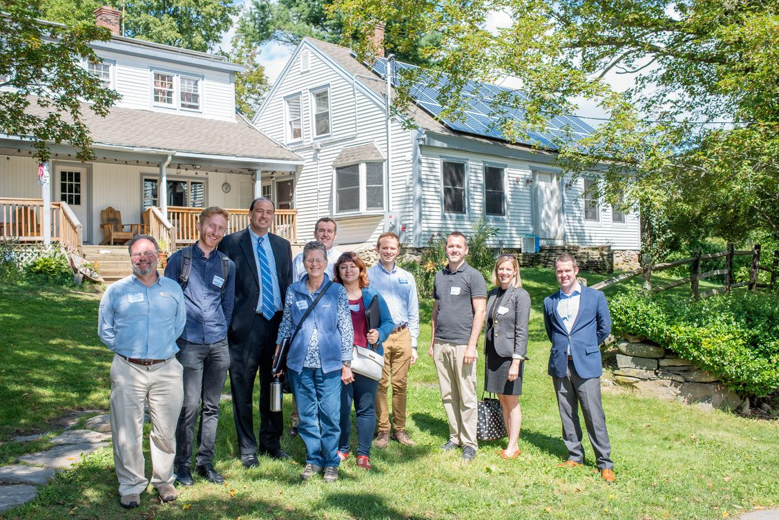 About a dozen individuals, dressed formally, standing in front of a farmhouse on a sunny day in the summer.