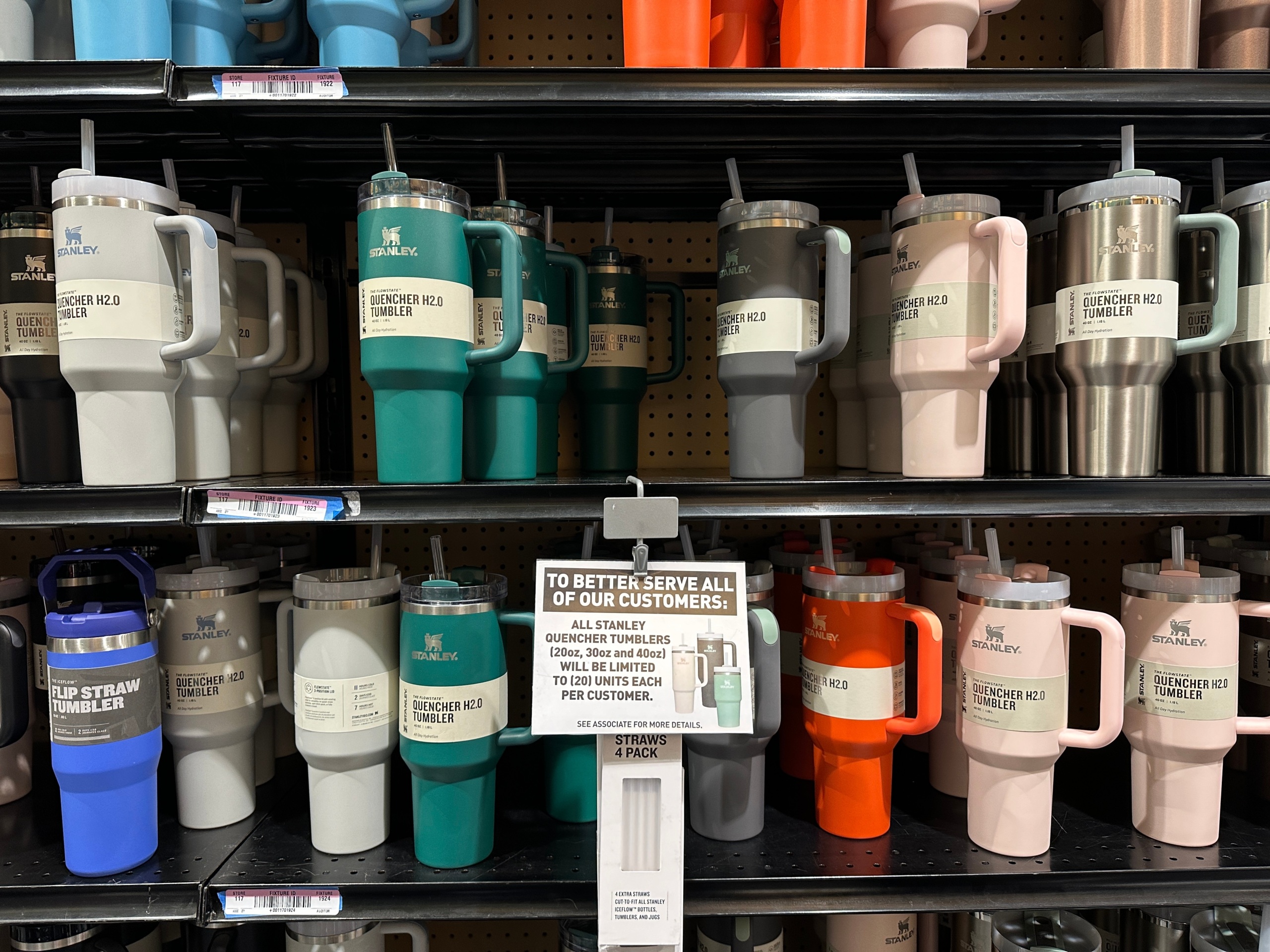 Stanley tumblers in a a variety of colors are for sale on a store shelf. A sign limits sales to 20 per customer.