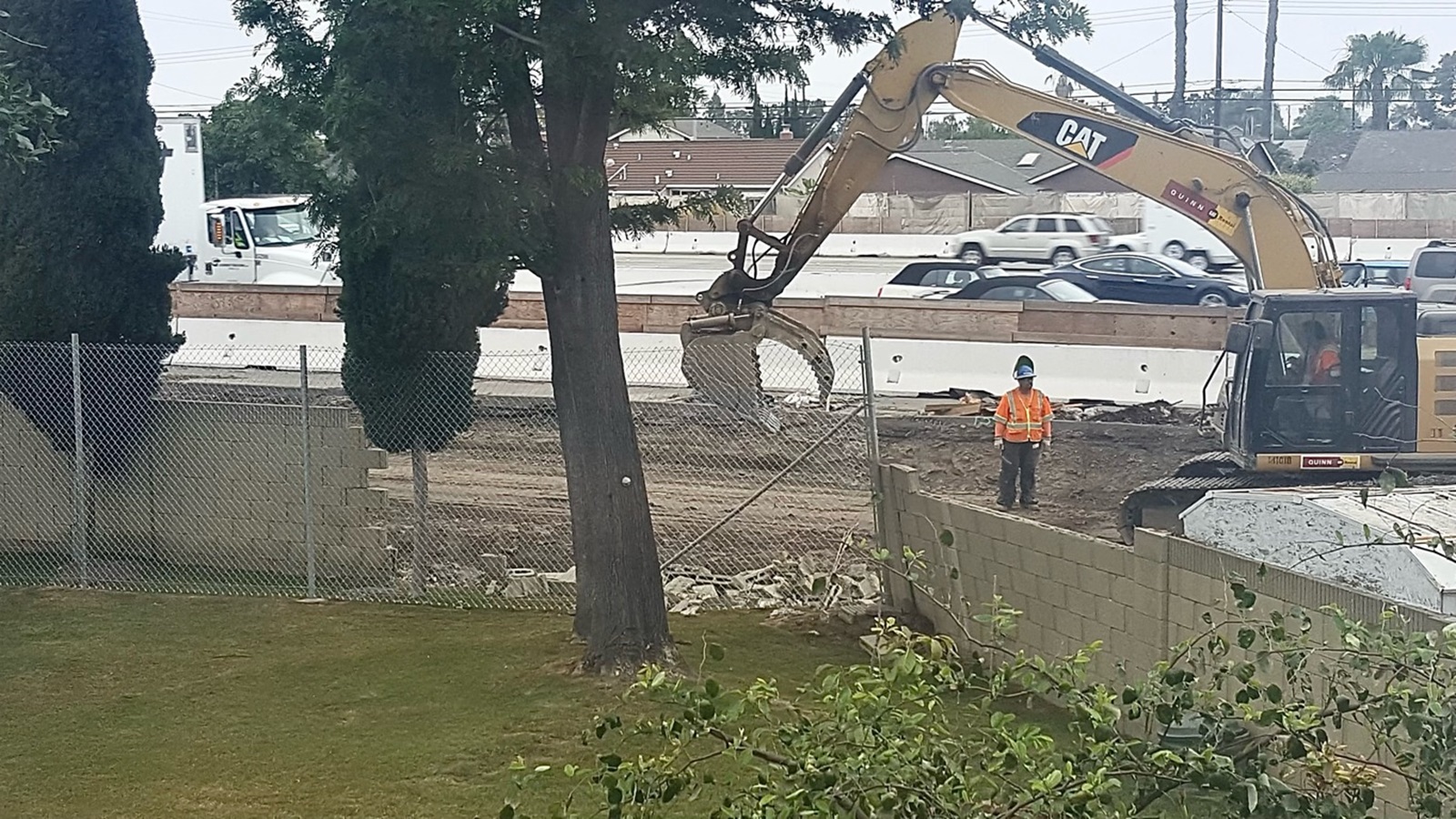 A backhoe and a construction worker at work between a busy freeway and a residential yard with a tree and green grass.
