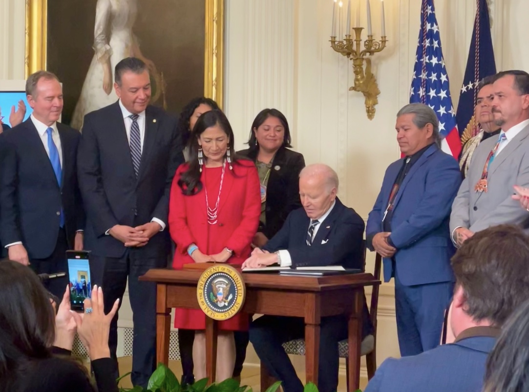 President Biden signing at a desk with Secretary Haaland, Senator Padilla, Senator Schiff, and indigenous leaders watching.