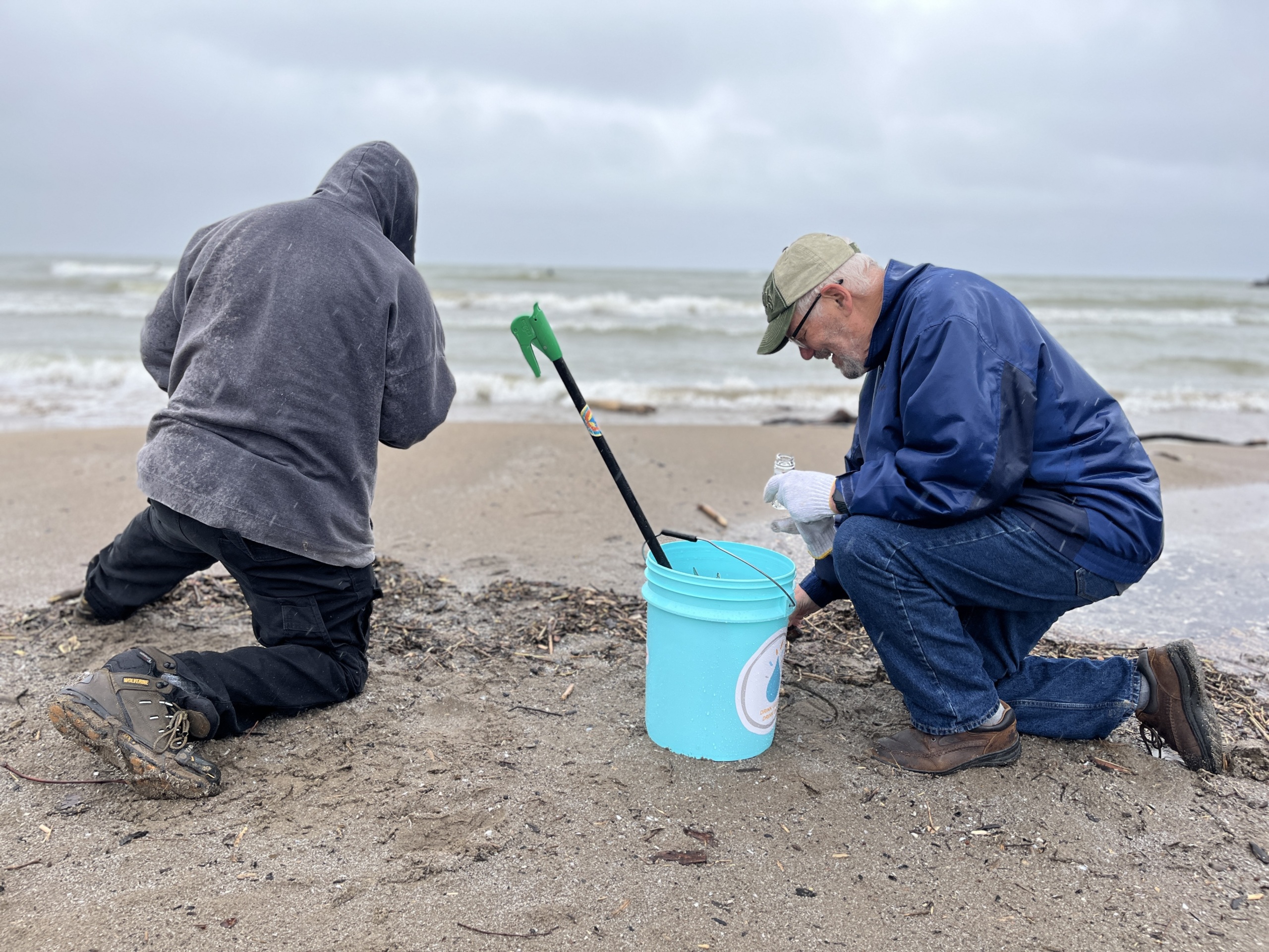 Two people on the beach on their knees with Lake Erie in the background looking for plastic pellets